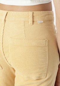 Pantaloni di corduroy gialli con righe verticali, dotati di una tasca posteriore e di un piccolo logo vicino alla vita. Tessuto testurizzato e design aderente.