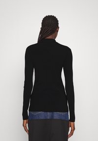 Svart långärmad ribbad tröja med krage, figursydd design och textilstruktur. Bärs över en denimskjorta, vilket ger ett lager-på-lager utseende.