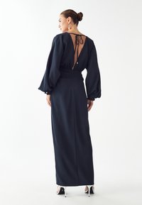 Námorníckomodrá šaty s vysokým pásom, dlhými nadýchanými rukávmi a odhaleným chrbtom zabezpečeným tenkým kravatom. Hladká, jemne tečúca látka siahajúca až po členky.