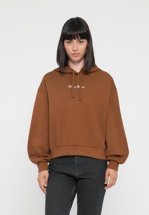 Brun hettegenser med en cropped design, snorhette, og ballongsleeves. Har hvit brodert tekst på brystet. Kombinert med mørke jeans.