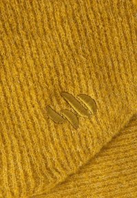 Geknoopt mosterdkleurig fabric met een textuur en subtiele ribbels. Heeft een geborduurd logo in een lichtere tint.