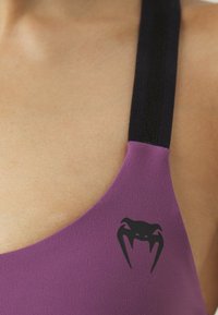 Vijolična športna modrc s črnim logotipom, širokimi črnimi naramnicami in gladko teksturo. Oblikovanje poudarja eleganten in prilegajoč obliko.