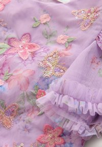 Tela lila con flores bordadas y mariposas en tonos rosa y verde, adornada con lentejuelas y detalles fruncidos en los bordes.