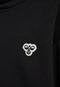 Schwarzer Stoff mit glatter Textur, der auf der unteren linken Seite ein weißes gesticktes Bienenlogo zeigt. Der Ausschnitt ist rund.