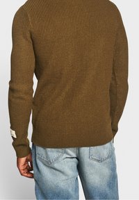 Man die een olijfgroene geribde trui en lichtblauwe spijkerbroek draagt, gezien van achteren tegen een effen achtergrond.