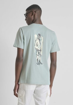Homme aux cheveux courts et lunettes de soleil portant un t-shirt bleu clair avec un dessin en noir et blanc d’un homme en short dans le dos.