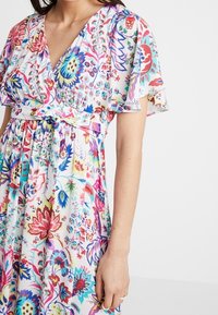 Blomstermønstret kjole i transparent stof med dyb V-hals, korte flæseærmer og knyttet talje. Farverne inkluderer pink, blå, grøn og gul.