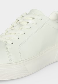Basket blanche basse avec lacets blancs, coutures visibles et semelle en caoutchouc texturée sur fond clair.