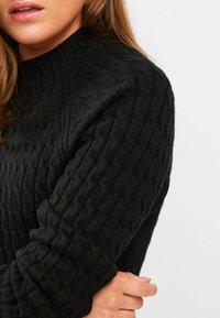Pull noir en tricot avec un design en câble texturé. Col montant et manches longues, montrant un gros plan du matériau.