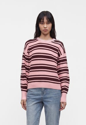 Ung kvinde iført en pink og mørkebrun stribet sweater og lyseblå jeans står mod en ensfarvet hvid baggrund.