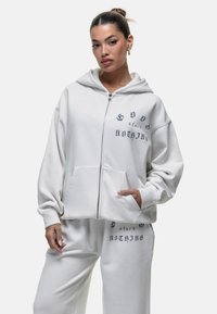 Felpa bianca con zip realizzata in tessuto morbido, dotata di una tasca frontale e testo stampato in blu navy sul petto sinistro e sulla schiena.