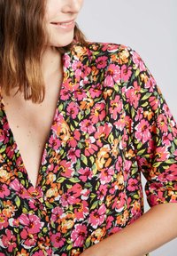 Blouse fleuri avec des fleurs roses, oranges et vertes vives sur un fond noir. Présente un décolleté plongeant et des manches courtes. Tissu lisse.
