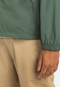 Grüne, leichte Jacke mit elastischen Bündchen, glatter Textur, seitlichen Taschen und kombiniert mit beigen Hosen. Das Material scheint wasserabweisend zu sein.