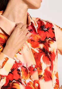 Frau richtet den Kragen eines beigen Hemdes mit großem rot-orangefarbenem Blumenmuster aus, das Knöpfe und lange Ärmel zeigt.