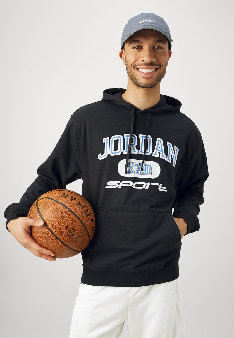 Sudadera negra con "JORDAN" y "sport" impresas en blanco y azul. Llevando pantalones cortos blancos, sosteniendo un balón de baloncesto. Gorra gris claro.