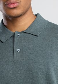 Polo en tela verde apagado, con un collar clásico, tapeta de dos botones y textura acanalada con detalles sutiles.