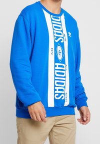 Sudadera azul de algodón con rayas verticales blancas que muestran el texto y logo "ADIDAS", cuello redondo y puños acanalados.
