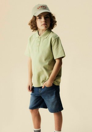 Garçon aux cheveux bouclés portant une casquette beige, un polo vert clair, un short en jean et des chaussettes rayées, debout avec une main dans la poche.