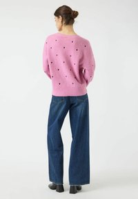 Rosa Strickpullover mit kleinen dunklen Herzmustern, kombiniert mit weit geschnittenen blauen Jeans und schwarzen High Heels. Einfarbiger Hintergrund.