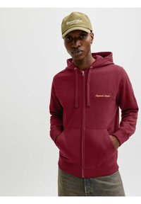 Sweat à capuche zippé couleur bordeaux avec poche kangourou, capuche à cordon de serrage et logo brodé. Associé à une casquette beige avec du texte blanc.