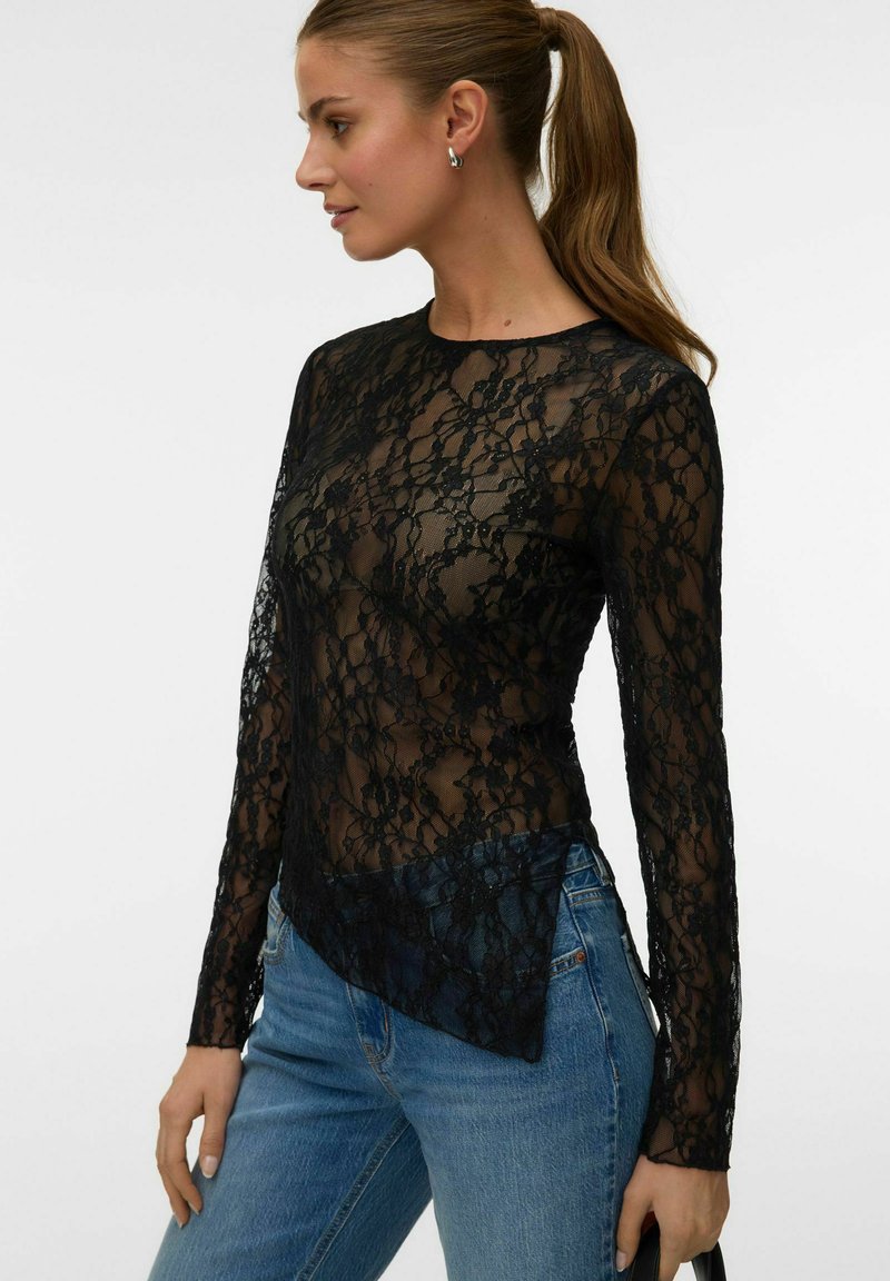 Femme aux cheveux bruns en queue de cheval portant un haut noir en dentelle florale à manches longues par-dessus une sous-couche noire et un jean bleu, tournée vers la gauche sur un fond uni.