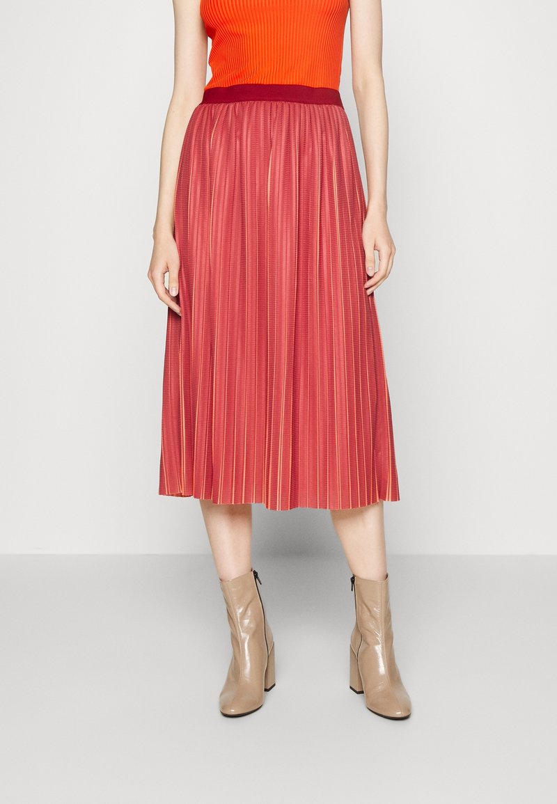 Jupe midi plissée rouge avec une taille contrastante foncée. Portée avec des bottines beiges, mettant en valeur une texture lisse et une silhouette fluide.