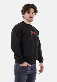 Maglione di cotone nero con maniche lunghe e collo a girocollo, caratterizzato da un logo grafico rosso sul petto. Indossato con jeans di denim blu.