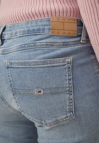 Vaqueros de mezclilla azul con una etiqueta de cuero marrón que dice "TOMMY JEANS". Presenta un pequeño parche de logo en colores rojo, blanco y azul en el bolsillo trasero.