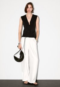 Donna che indossa un top nero senza maniche, pantaloni bianchi larghi, bracciali d'argento, con in mano una borsa nera a forma di mezzaluna, in piedi di fronte a un muro bianco.