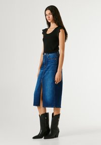 Naine, kes kannab musta varrukateta toppi, sinisest denimist midi-seelikut eeslõhikuga ning musti keskmise sääreni ulatuvaid teravate ninadega saapaid, seisab valge tausta ees.