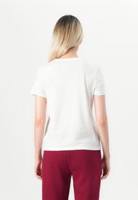 Persona con lunghi capelli biondi che indossa una semplice maglietta bianca a maniche corte e pantaloni rossi, di spalle su uno sfondo bianco.