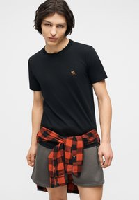 Νέος με καστανά μαλλιά, φοράει μαύρο T-shirt με μικρό λογότυπο ελαφιού, γκρίζα σορτς και κόκκινο-μαύρο καρό πουκάμισο δεμένο γύρω από τη μέση.