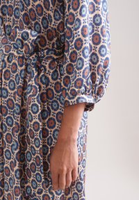 Kleid aus floral gemustertem Stoff in hellpfirsich, mit blauen und orangen Blumen. Der Ärmel ist gerafft und mit einem Bündchen versehen, einschließlich eines selbstgebundenen Gürtels.