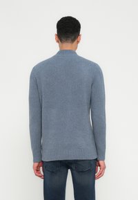 Egy férfi kék kötött pulóverben és sötét farmernadrágban, háttal a kamerának, egy sima fehér háttér előtt.