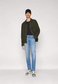 Jachetă verde măsliniu cu nasturi și buzunar aplicat, tricou negru, blugi albaștri deschis și adidași multicolori. O șapcă neagră completează look-ul.