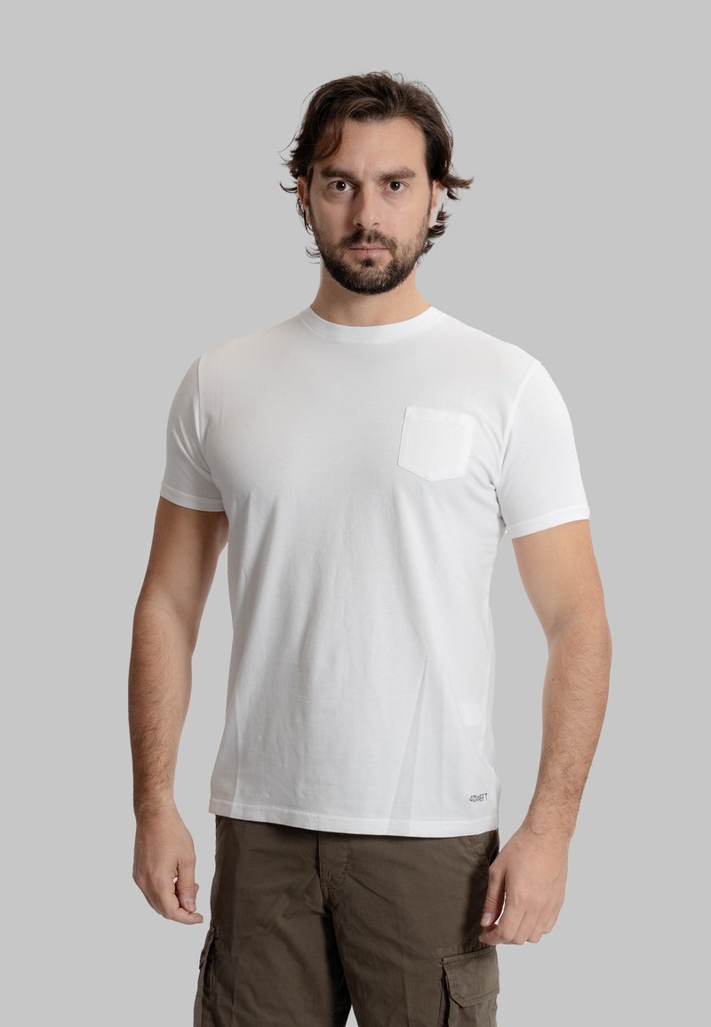 Maglietta bianca in cotone con collo rotondo e maniche corte, con taschino sul petto e orlo dritto. Il modello indossa dei pantaloni corto color khaki.