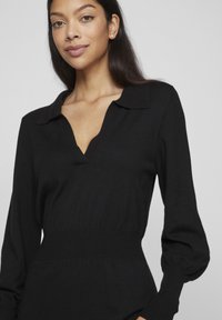 Juodas megztinis ilgomis rankovėmis su V formos iškirpte ir apykakle, pasižymintis siauru liemeniu ir pūstomis rankovėmis, lygios tekstūros be matomų raštų.