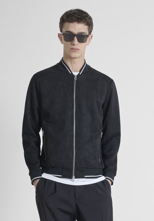 Chaqueta bomber de ante negra con cuello y puños acanalados, detalles en blanco, cierre de cremallera y bolsillos laterales. Llevada sobre una camiseta blanca.