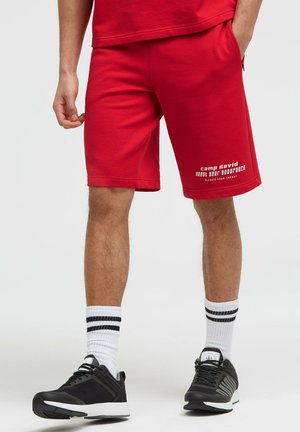 Mann trägt rote Sportshorts mit weißem Text am Bein, weiße Socken mit schwarzen Streifen und schwarze Sportschuhe, steht mit einer Hand in der Hosentasche.