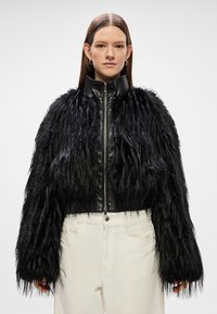 Casaco curto de pelúcia sintética negra com colarinho e mangas em couro liso. Apresenta um fecho frontal e detalhes em franjas na barra.
