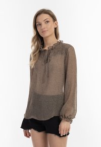 Gennemsigtig brun bluse med blomstermønster, flæset halsudskæring, bindelukning og elastiske manchetter, sammen med mørke shorts.