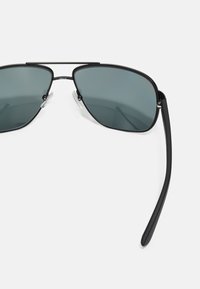 Ochelari de soare aviator cu lentile gri, cadru metalic negru și bară superioară distinctivă. Dispune de tampoane nazale transparente și brațe subțiri negre.