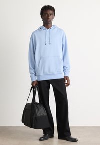 Sweatshirt bleu deschis cu glugă și buzunar frontal, purtat cu pantaloni negri largi și o geantă de duffel neagră. Pantofi din piele mată.