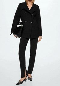 Manteau noir croisé avec de grands boutons, associé à un pantalon noir ajusté avec une fente sur le côté, et des chaussures à petits talons noires ornées.