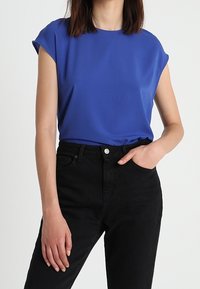 Modrý top s krátkým rukávem a hladkou texturou, kombinovaný s černými džínami s vysokým pasem. Top má volnější střih a kulatý výstřih.