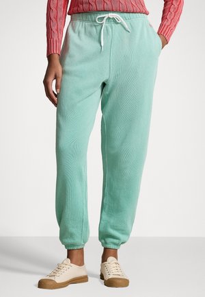 Vrouw draagt mintgroene joggingbroek met een wit koordje, gecombineerd met een roze kabelgebreide trui en beige sneakers met bruine zolen.