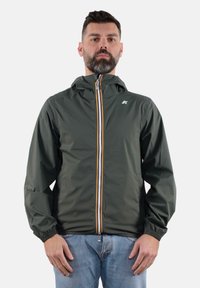 Giacca impermeabile verde con zip frontale, polsini elasticizzati e cappuccio. Presenta un logo bianco e dettagli della zip gialli e arancioni a contrasto.