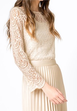 Vrouw draagt een crème kleurige, lange mouwen kanten top gecombineerd met een hooggetailleerde, geplooide crème rok, met details van arm en torso zichtbaar.
