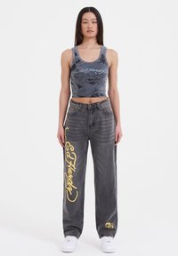 Grå cropped tank top med sort drageprint, parret med højtaljede grå jeans med gule Ed Hardy-tekster og grafiske detaljer.