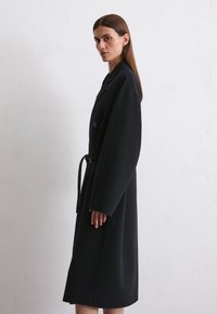 Black wool coat with a long, straight silhouette. Features wide sleeves, a tied waist, and two buttons on the front. Smooth texture.